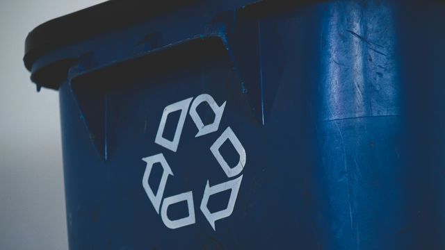 A close-up photo of a blue recycling bin, with the triangular recycling symbol printed in white on the front. 